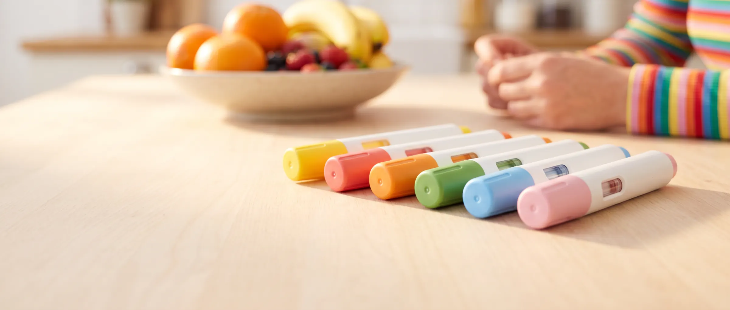 Six plain injection pens with coloured caps lying side by side on a sunny table