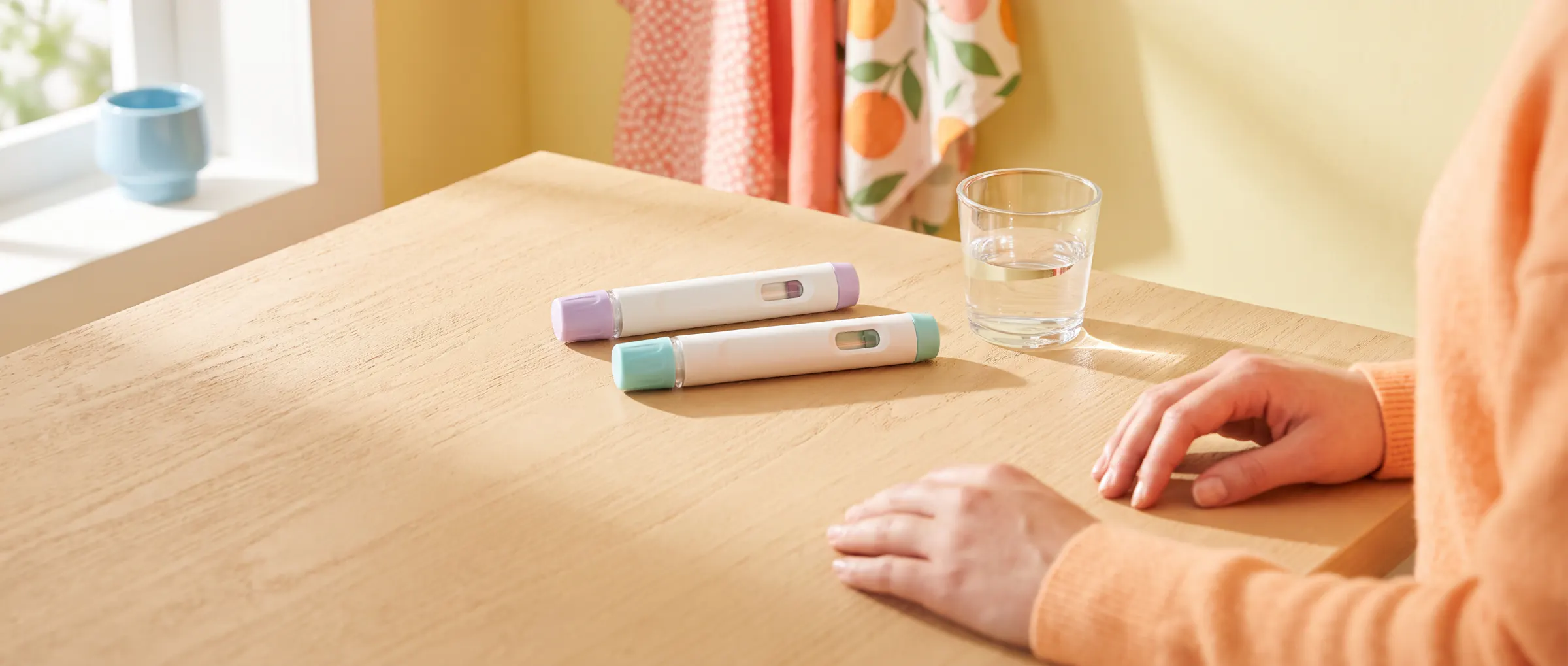 Two plain injection pens, one lilac and one teal, side by side on a sunny table beside a glass of water