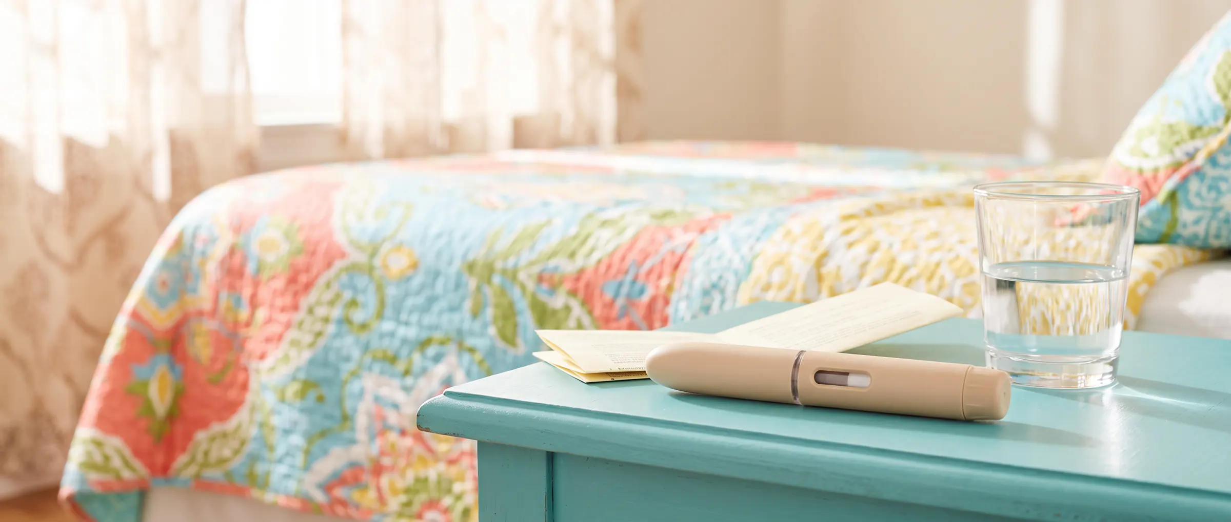 A plain injection pen, a folded leaflet and a glass of water on a bright bedside table
